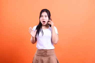 Asya 'lı bir kadın ararken şok oluyor. O, turuncu bir arka planda izole edilmiş beyaz bir gömlek ve kahverengi bir etek giyiyor. Ağzı açık ve şaşırmak için ellerini kaldırıyor..