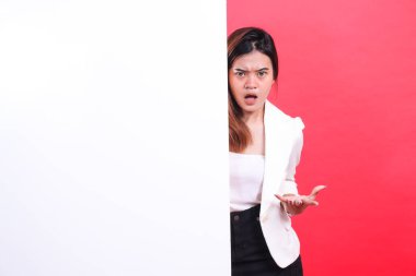 Şaşırmış bir ifadeyle beyaz bir tahtanın arkasından dışarı bakan eli açık ve kırmızı arka planda izole bir şekilde kameraya bakan bir kadın.