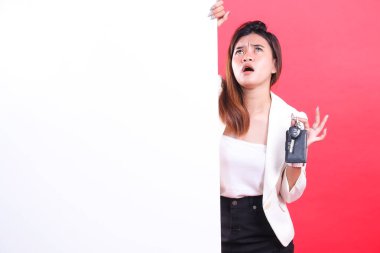 Bir kadın elinde siyah bir anahtar ve beyaz bir tabela tutuyor, yukarı bakıyor, beyaz bir bluz ve ceket giyiyor, kırmızı bir arka planda izole edilmiş.