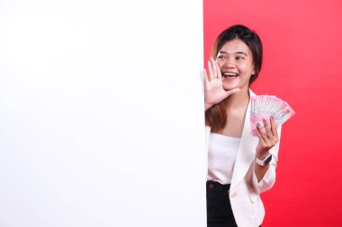 Beyaz ceketli bir kadın elinde para, kırmızı arka planda izole edilmiş beyaz bir pankartın yanında elini sallıyor.