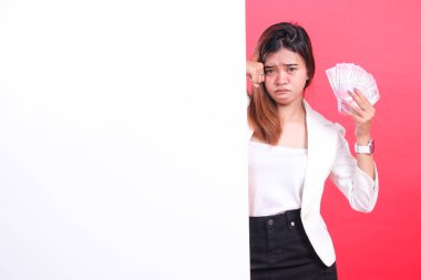 Bir yığın parası olan bir kadın elinde tutuyor ve üzgün görünüyor, beyaz bir tahtanın arkasında duruyor, kırmızı bir arka planda soyutlanmış.