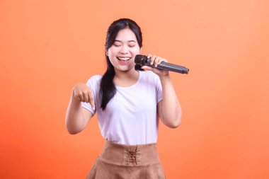 Neşeli genç bir kadın mikrofona şarkı söylüyor ve aşağıya doğru gülümsüyor, turuncu arka planda soyutlanmış..