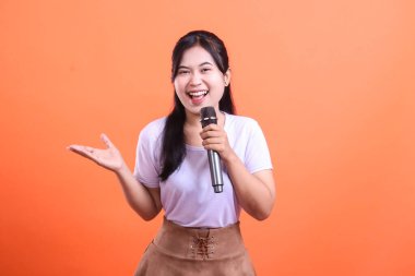 Elinde mikrofon tutan neşeli bir kadın. Turuncu arka planda mutlu ve kendinden emin bir şekilde el kol hareketi yapıyor.