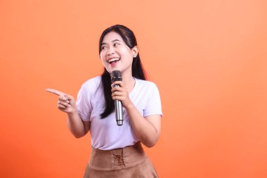 Neşeli bir kadın, elinde bir mikrofon tutarken ve parmağıyla el kol hareketi yaparken, coşku ve nişanı iletirken ortaya çıkıyor. turuncu arkaplanda izole