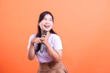 Genç bir kadın bir mikrofon tutuyor ve anlamlı bir gülümseme ve jestle şarkı söylüyor, kendinden emin ve mutlu görünüyor. turuncu arkaplanda izole