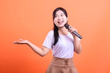Asyalı bir kadın mikrofonu tutarken gülümsüyor ve diğer eliyle sunuyor. Turuncu arka planda izole edilmiş sevinç ve heyecanını dile getiriyor.