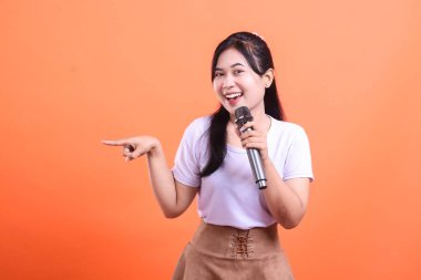 Elinde mikrofon tutan ve yan tarafı gösteren gülümseyen genç bir kadın sunum ya da eğlence konsepti için mükemmel. turuncu arkaplanda izole