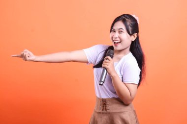 Gülümseyen genç bir kadın elinde bir mikrofon tutarken enerjik bir şekilde işaret eder. Heyecanlı ve turuncu arka planda izole edilmiş bir şekilde sunumuna hazır görünüyor.