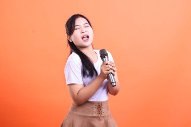 Genç bir kadın tutkuyla mikrofona şarkı söylüyor, gözleri kapalı, tüm kalbiyle müziğin tadını çıkarıyor, turuncu arka planda izole bir şekilde.