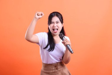 Genç, mutlu bir kadın heyecanla mikrofona şarkı söylüyor ve yumruğunu kaldırıyor. Turuncu arka planda tutkusunu ve zevkini gösteriyor.