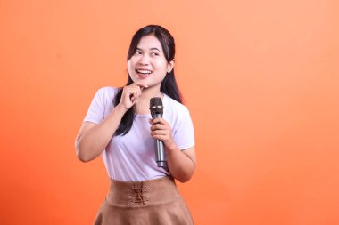 Bir kadın mikrofonu incelikle tutuyor, muhtemelen bir performansa hazırlanıyor, yüzünde bir gülümseme ve çenesinde de parmağı var. turuncu arkaplanda izole