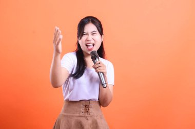 Genç ve anlamlı bir kadın mikrofona tutkuyla şarkı söylüyor. Kolu dinamik bir jestle kaldırılmış, turuncu arka planda izole edilmiş..