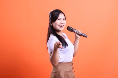 Gülümseyen Asyalı bir kadın bir mikrofon tutuyor ve parmağını öne doğru uzatıyor, turuncu arka planda izole edilmiş neşeli bir yüz ifadesi sergiliyor.