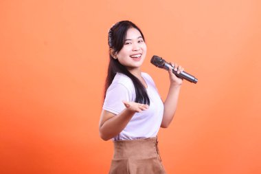 Neşeli genç bir kadın mikrofon tutuyor ve şarkı söylüyor, geniş bir şekilde gülümsüyor ve turuncu arka planda izleyicilerle iletişim kuruyor.