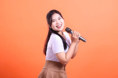 Mikrofon tutan ve mutlu bir şekilde şarkı söyleyen genç bir kadın, müziğe olan tutkusunu ışıltılı bir gülümsemeyle ifade ediyor. turuncu arkaplanda izole