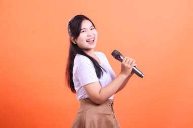 Kameraya bakarken mikrofona şarkı söylemekten zevk alan neşeli genç bir kadın, çok mutlu ve anlamlı görünüyor. turuncu arkaplanda izole