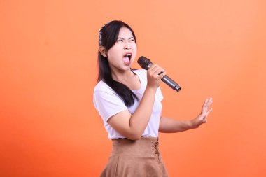 Tutkulu bir şekilde mikrofona şarkı söyleyen genç bir kadın yoğun bir duygu ve ağzı açık, elleri havada, müziğe olan tutkusunu gösteriyor. turuncu arkaplanda izole