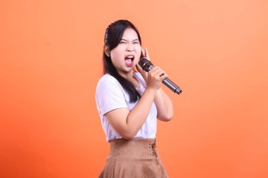 Genç, enerjik bir kadın mikrofona tutkuyla şarkı söylüyor. Duygularını turuncu arka planda izole edilmiş güçlü bir performansla ifade ediyor..