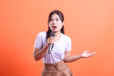 Genç bir kadın, elinde mikrofonla, açık ağızlı ve turuncu arka planda soyutlanmış bir şekilde şarkı söylüyor ve el kol hareketi yapıyor.