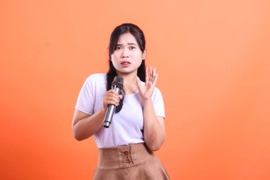 Mikrofonlu endişeli Asyalı kadın, turuncu arka planda izole edilmiş elleriyle bir jest yapıyor..