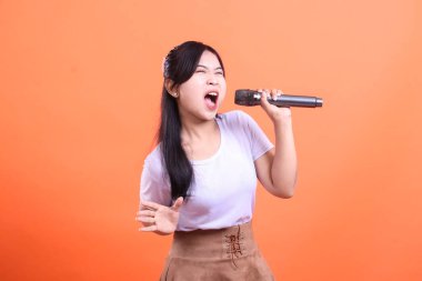 Tutkulu bir ifadeyle elinde mikrofonla şarkı söyleyen Asyalı genç bir kadın ağzı açık, turuncu arka planda izole bir şekilde şarkı söylüyor.