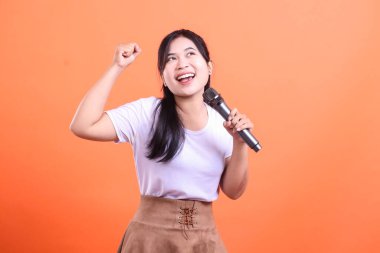 Genç bir kadın mikrofonu tutarken sevinçle yumruğunu kaldırıyor, sanki turuncu arka planda büyük bir performansı kutluyor ya da kazanıyormuş gibi..