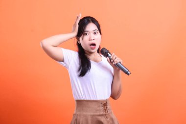 Şaşırmış bir yüzü olan bir kadın mikrofona şarkı söylüyor. Bir eli başının üstünde ve ağzı açık, turuncu arka planda izole bir şekilde.