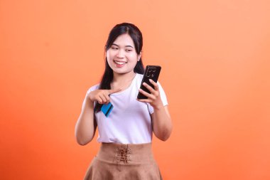 Genç bir kadın cep telefonunu kullanırken gülümsüyor ve kredi kartını işaret ederek, turuncu arka planda izole edilmiş online alım-satım veya işlemleri öneriyor.