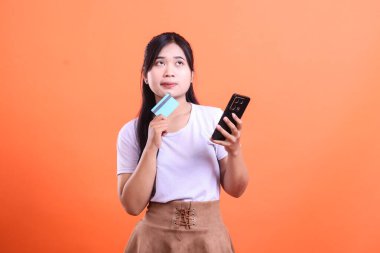 Genç bir Asyalı kadın elinde bir kredi kartı ve bir cihaz telefonu tutarken yukarı bakıp online alışveriş fikirleri hakkında düşünüyor. turuncu arkaplanda izole