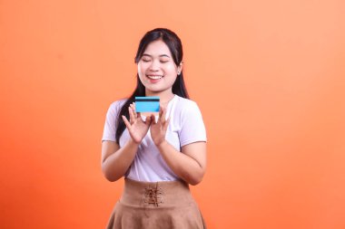 Gülümseyen genç bir kadın iki eliyle kredi kartı tutuyor, yeni finansal olanaklardan memnun, turuncu arka planda izole edilmiş..