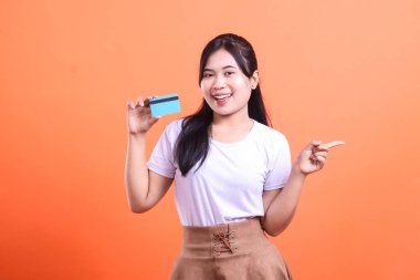 Elinde kredi kartıyla gülümseyen bir kadın, parmağını sağa gösteriyor, kameraya bakıyor ve turuncu arka planda izole edilmiş mutlu bir yüz ifadesi gösteriyor.