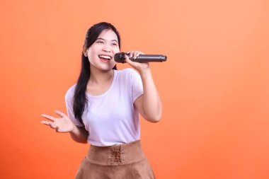 Mikrofon tutan ve mutlulukla şarkı söyleyen bir kadın, çok etkileyici görünür ve performans gösterirken elleriyle işaret eder, turuncu arka planda izole bir şekilde.