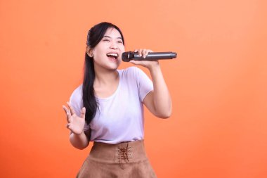 Genç bir kadın, neşeli bir şekilde mikrofona şarkı söylüyor. Parlak bir gülümsemeyle müziğe ve gösteriye olan tutkusunu ifade ediyor. turuncu arkaplanda izole