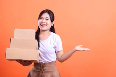 Elinde karton kutularla müşterilere sipariş vermeye hazır gülümseyen genç bir kadın, turuncu arka planda izole edilmiş müşterilerine iyi hizmet vermekten mutluluk duyar.