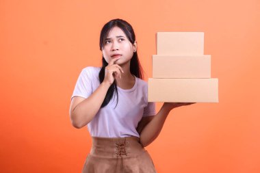 Genç Asyalı bir kadın elinde bir yığın karton kutu tutarak düşünceli bir şekilde bakıyor, bir sonraki adımı ya da teslimat kararını düşünüyor, turuncu arka planda izole edilmiş..