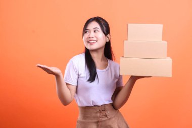 Genç bir Asyalı kadın elinde gülümsemeli üç karton kutu tutuyor ve diğer eliyle turuncu arka planda izole bir şekilde sunum yapıyor..