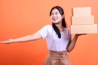 Genç Asyalı bir kadın bir elinde karton kutular, diğer elinde ise gülümseyen ve turuncu arka planda izole edilmiş tarafa bakan bir sunum yapıyor.