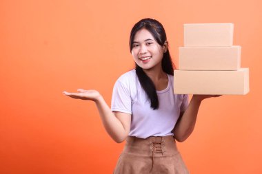 Elinde üç karton kutu tutan genç bir kadın diğer eliyle turuncu arka planda izole edilmiş bir şey sunuyor..