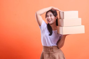 Genç bir kadın üç karton kutu taşıyor, mutlu görünüyor ve siparişi vermeye hazır, turuncu arka planda izole edilmiş.