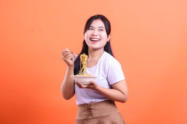 Kocaman gülümseyen, elinde bir kase erişteyle lezzetli yemeğinin tadını çıkarmaya hazır neşeli bir genç kadın. turuncu arkaplanda izole