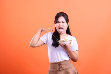 Genç Asyalı kadın elindeki eriştenin tadına baktıktan sonra suratını asıyor. Dilini uzatır ve elini kaldırır, turuncu arka planda izole bir şekilde.