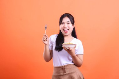 Parlak bir gülümsemesi olan genç bir kadın elinde bir kase makarna ve çatalla yemeğinin tadını çıkarmaya hazır. turuncu arkaplanda izole