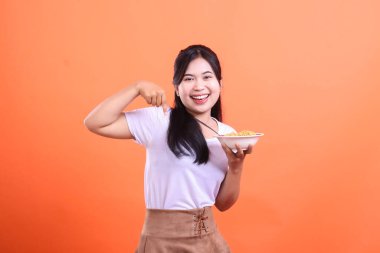 Genç ve mutlu bir kadın elinde bir kase erişteyi işaret ediyor, gülümsüyor ve neşeli görünüyor, turuncu arka planda soyutlanmış..