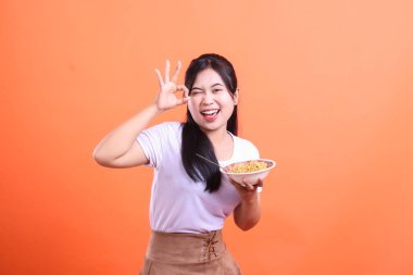 Neşeli bir Asyalı kadın göz kırpıyor ve elinde bir kase şehriye, geniş bir gülümsemeyle turuncu arka planda izole edilmiş zevk ve memnuniyeti gösteren 