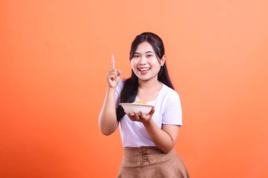 Gülümseyen genç bir kadın elinde bir kase erişteyle işaret parmağıyla turuncu arka planda mutlu ve mutlu görünüyor.