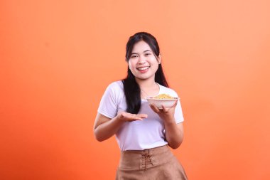 Neşeli genç bir kadın elinde lezzetli bir kase erişte tutuyor, bir jest ile sunuyor, yemeye hazır. turuncu arkaplanda izole