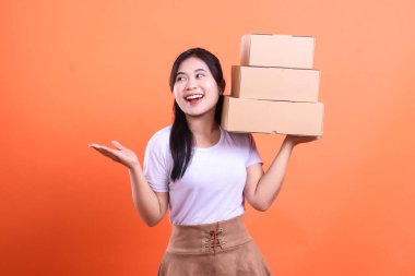 Gülümseyen genç bir kadın, turuncu arka planda izole edilmiş neşeli ve verimli bir teslimat ya da alışveriş deneyimi sergileyen üç karton kutu destesini elinde tutuyor.