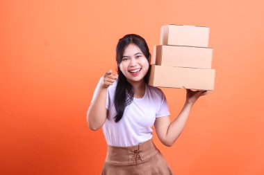 Kutuları tutan, gülümseyen bir yüzle kameraya bakan, turuncu arka planda izole edilmiş neşeli genç bir kadın.
