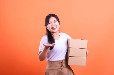 Karton kutuları olan neşeli bir teslimatçı kadın, gülümsüyor ve eli açık sunuyor. turuncu arkaplanda izole