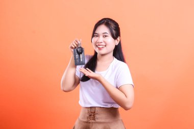 Gülümseyen Asyalı kadın araba anahtarlarını tutuyor ve sunuyor, yeni alımı veya başarıyı kutluyor, turuncu arka planda izole edilmiş..